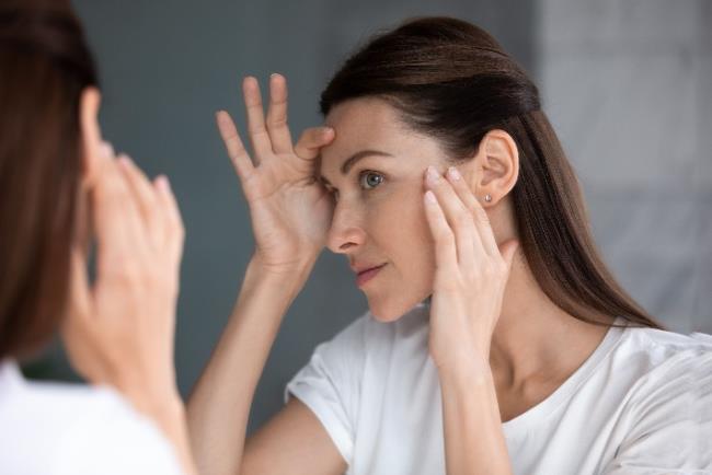 אישה בוחנת את פניה במראה כדי להחליט איזה טיפול אסתטי עליה לבצע כדי לשפר את מראה הפנים ומרקם העור